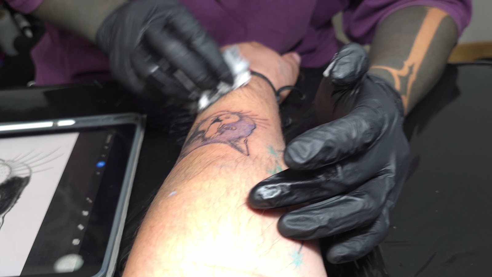 Manos con guantes negros sostienen una máquina de tatuar sobre un antebrazo con el dibujo ya calcado en violeta; al lado, una tableta con el boceto en pantalla.