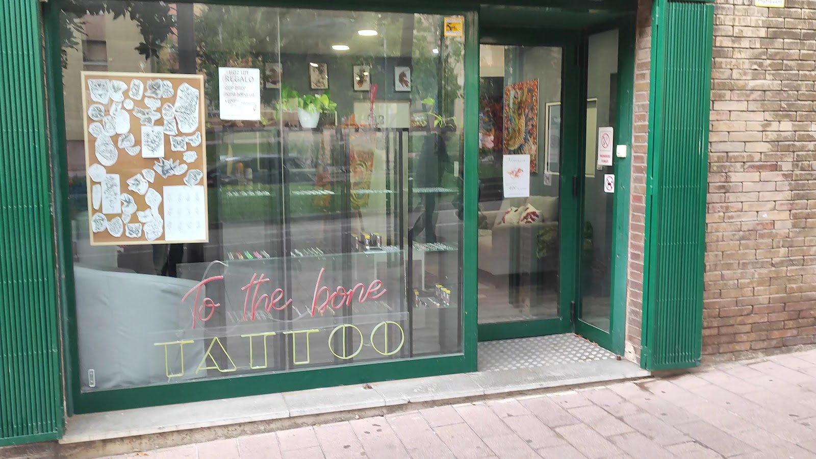 Escaparate con marco verde: rótulo de neón rosa con la palabra To the bone sobre las letras doradas y huecas de TATTOO, un corcho lleno de láminas de dibujos a la izquierda y el sofá y las plantas del interior al otro lado del cristal.